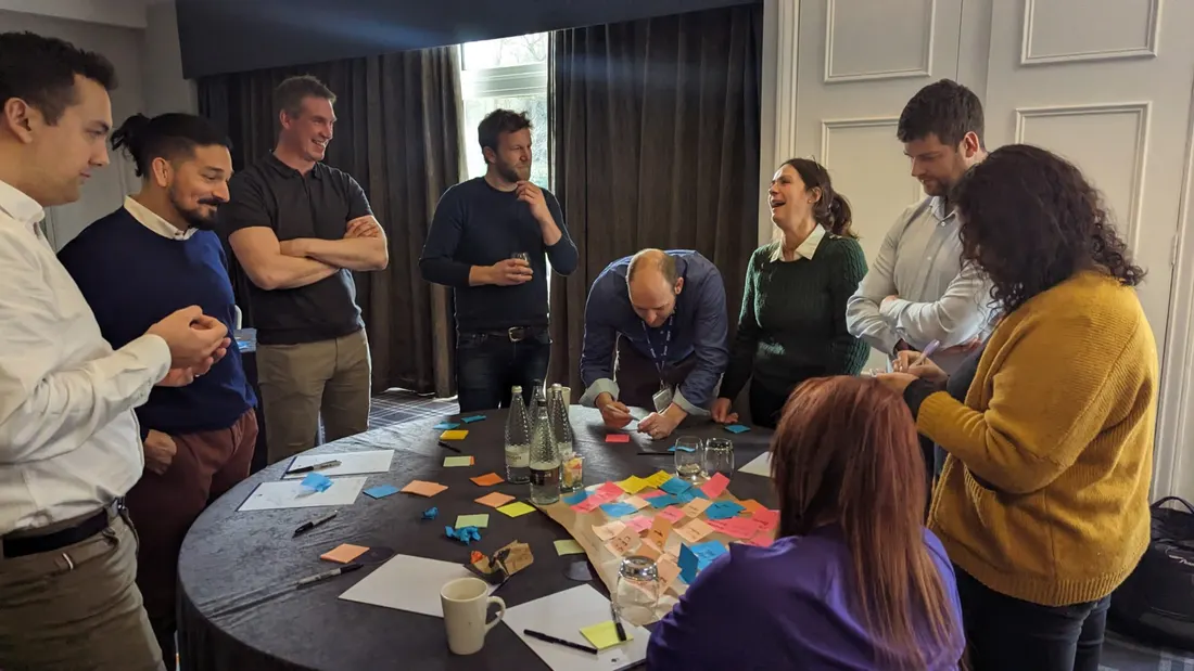 Une équipe debout autour d'une table ronde couverte de papillons adhésifs pendant un atelier, plusieurs personnes en train de rire