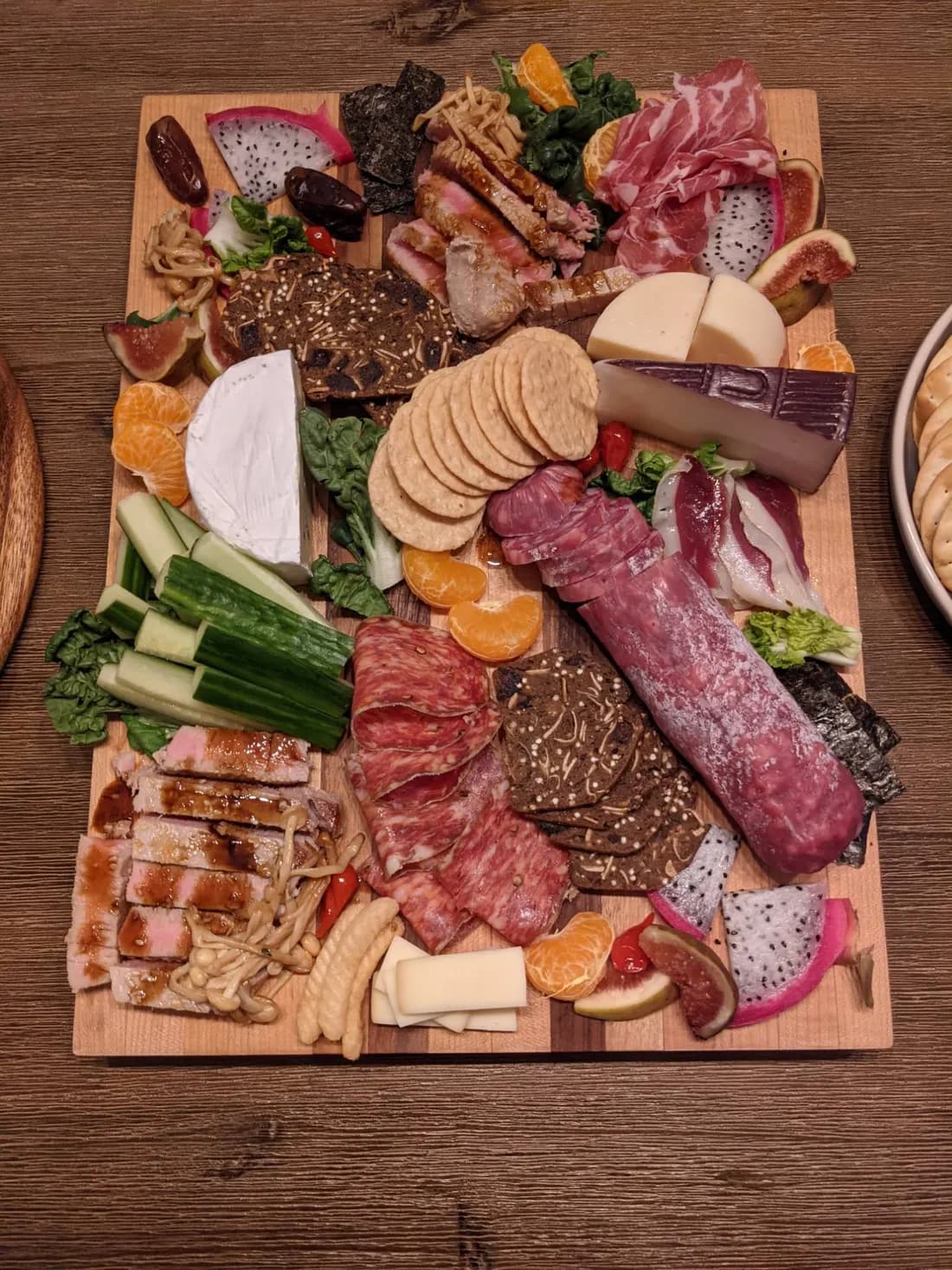 Une planche de partage avec du thon saisi glacé au soya et des champignons enoki, à côté de charcuteries, de fromages, de figues, de fruit du dragon, de clémentines et de nori